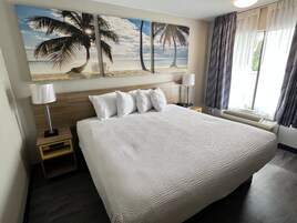 Habitación de hotel con una cama grande, una mesita de noche, una lámpara, un cuadro con una playa y una ventana con cortinas.