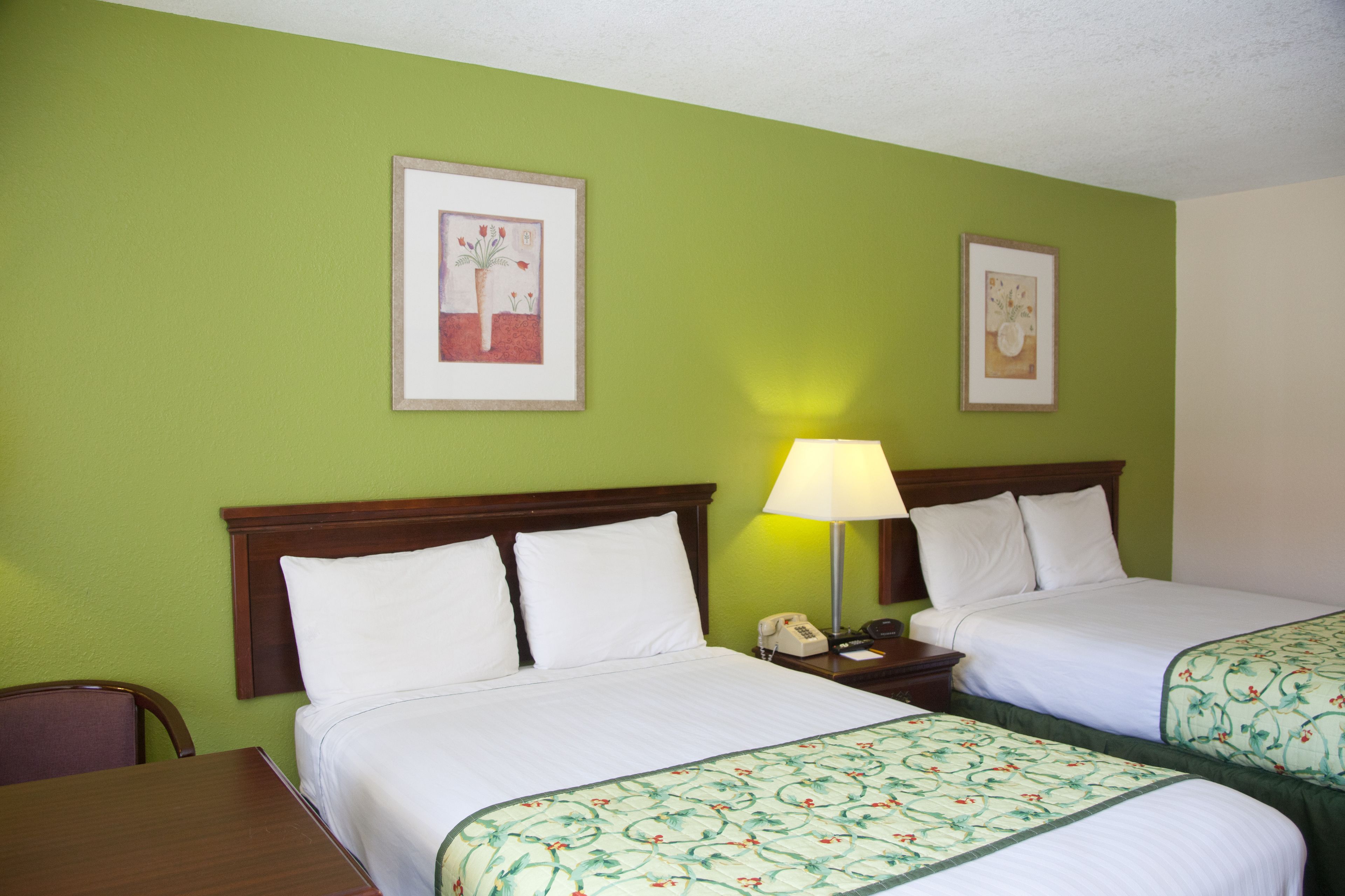 A hotel room with two beds, green walls, framed floral artwork, a bedside table with a lamp, and a wooden chair.