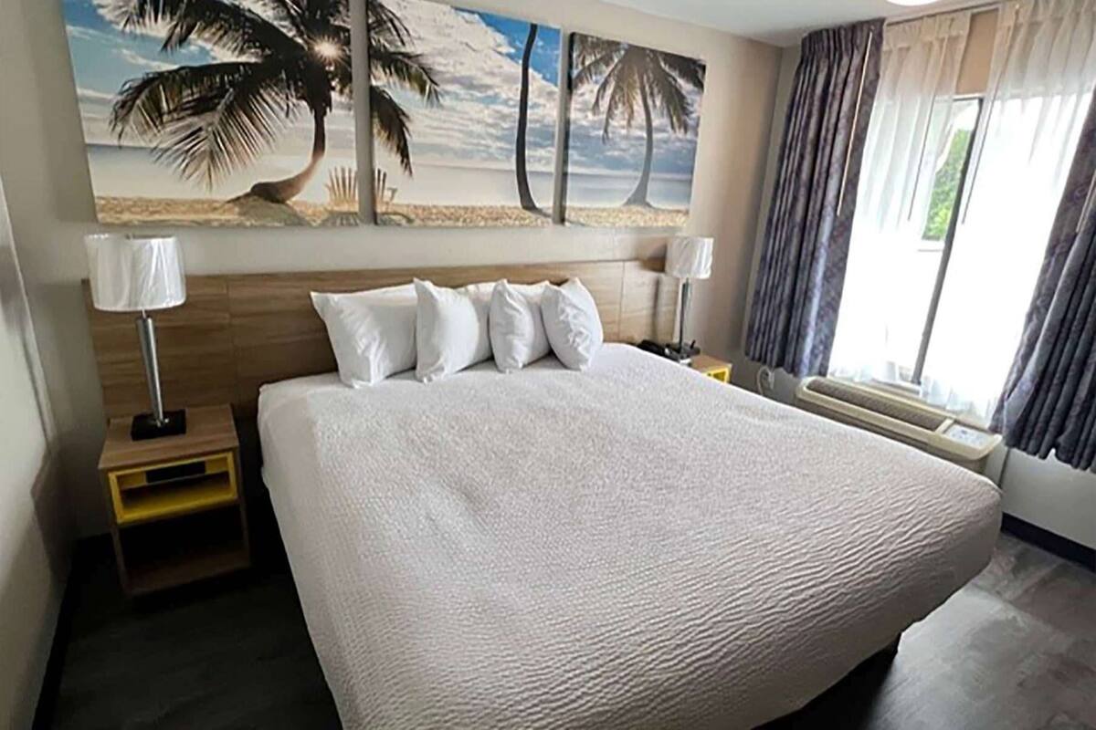 Quarto de hotel com uma cama grande, duas mesas de cabeceira, um criado-mudo, uma janela com cortinas e um quadro com tema de praia na parede.