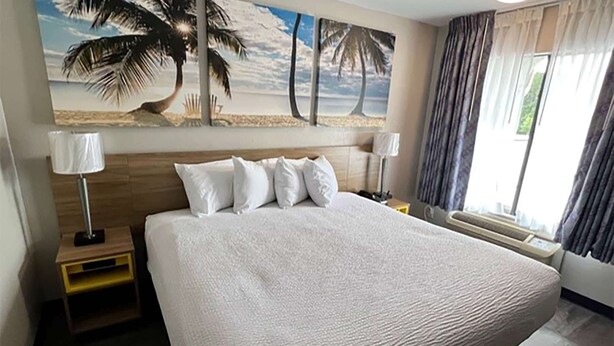 A hotel room with a large bed, two bedside tables, a nightstand, a window with curtains, and a beach-themed wall mural. - Days Inn by Wyndham DeFuniak Springs (De Funiak Springs)