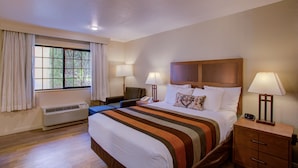 Ein Hotelzimmer mit einem großen Bett, zwei Nachttischen mit Lampen, einem Sessel, einem Schreibtisch und einem fenster mit Vorhängen.
