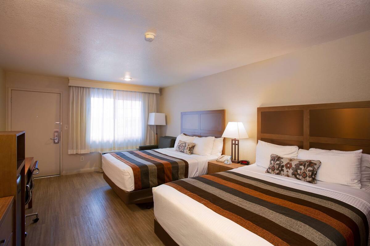 A hotel room with two beds, a desk, a chair, a lamp, and a window with curtains.