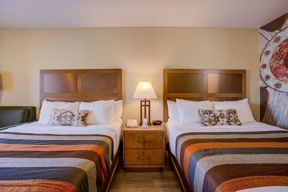 A hotel room with two beds, a nightstand with a lamp, a phone, and a small drawer.