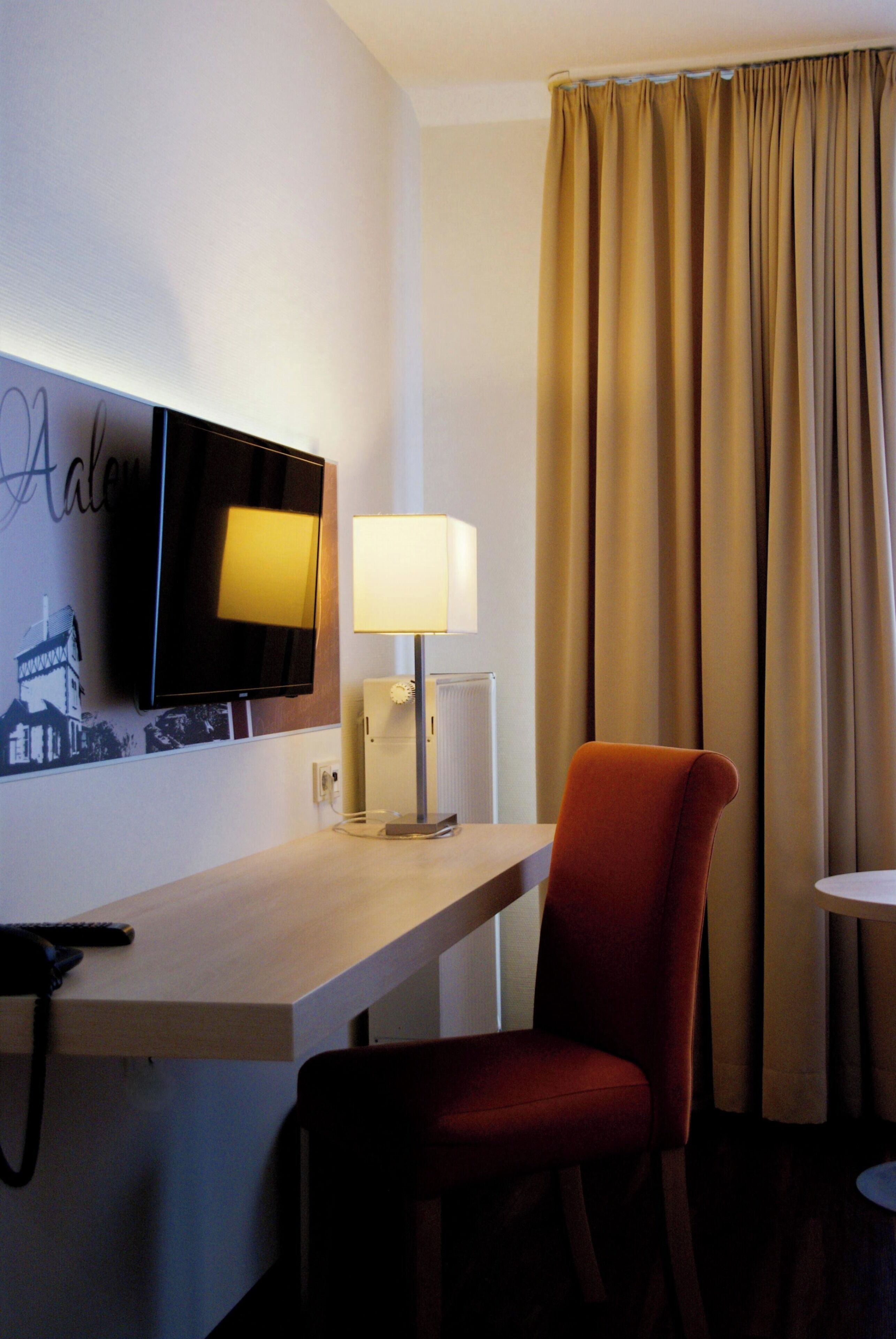 Habitación de hotel con televisión de pantalla plana montada en la pared, una mesita de noche con lámpara, una silla roja y una ventana con cortinas.