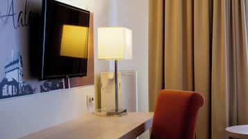 Habitación de hotel con televisión de pantalla plana montada en la pared, una mesita de noche con lámpara, una silla roja y una ventana con cortinas.