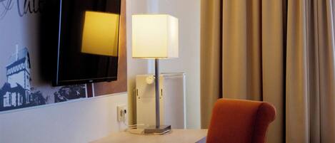 Habitación de hotel con televisión de pantalla plana montada en la pared, una mesita de noche con lámpara, una silla roja y una ventana con cortinas.