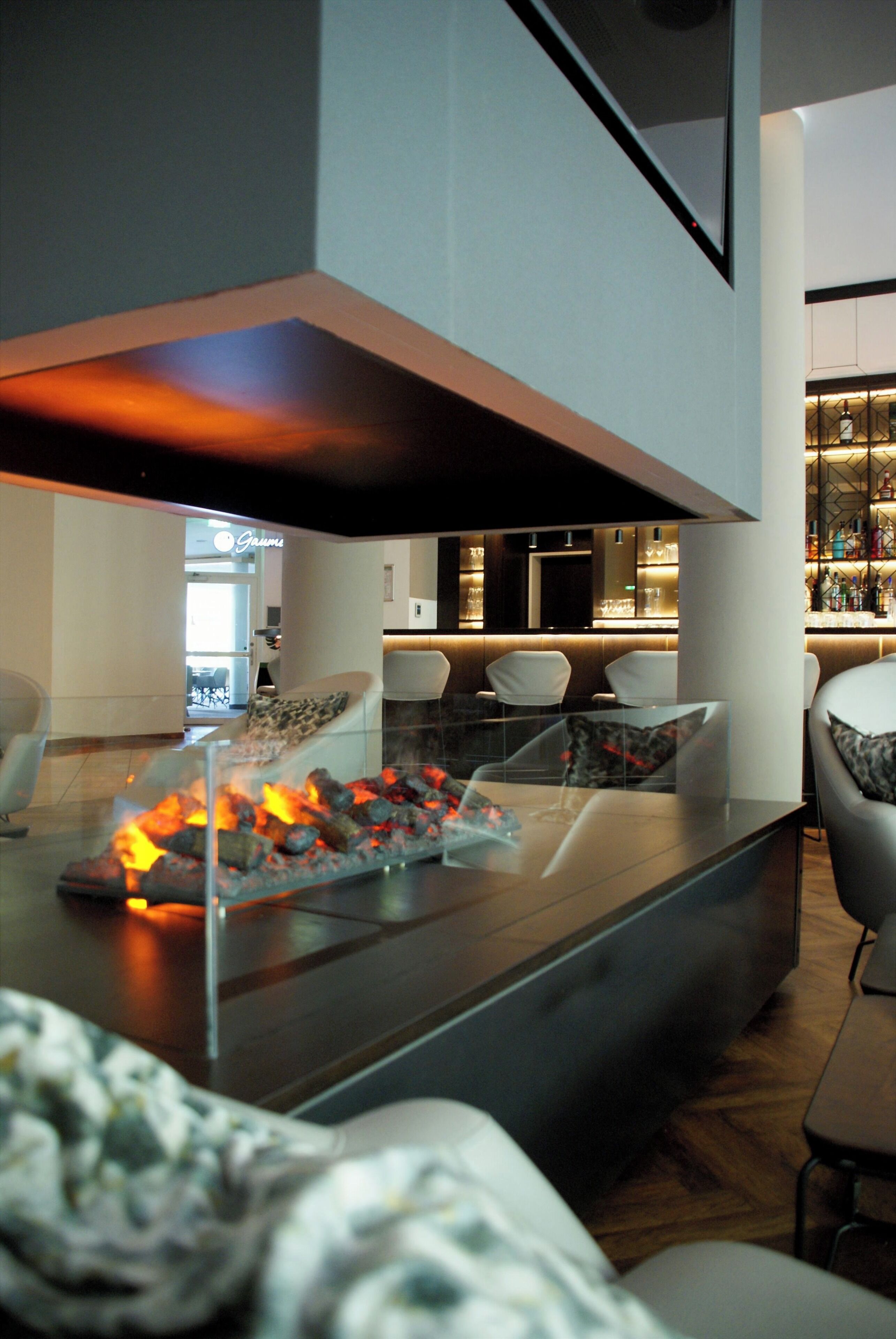 Ein modernes Interieur mit einem Tisch mit Glasplatte und Feuerstelle für Holzkohle, einer Sitzecke mit Sesseln und einer Bar mit Flaschenregalen.