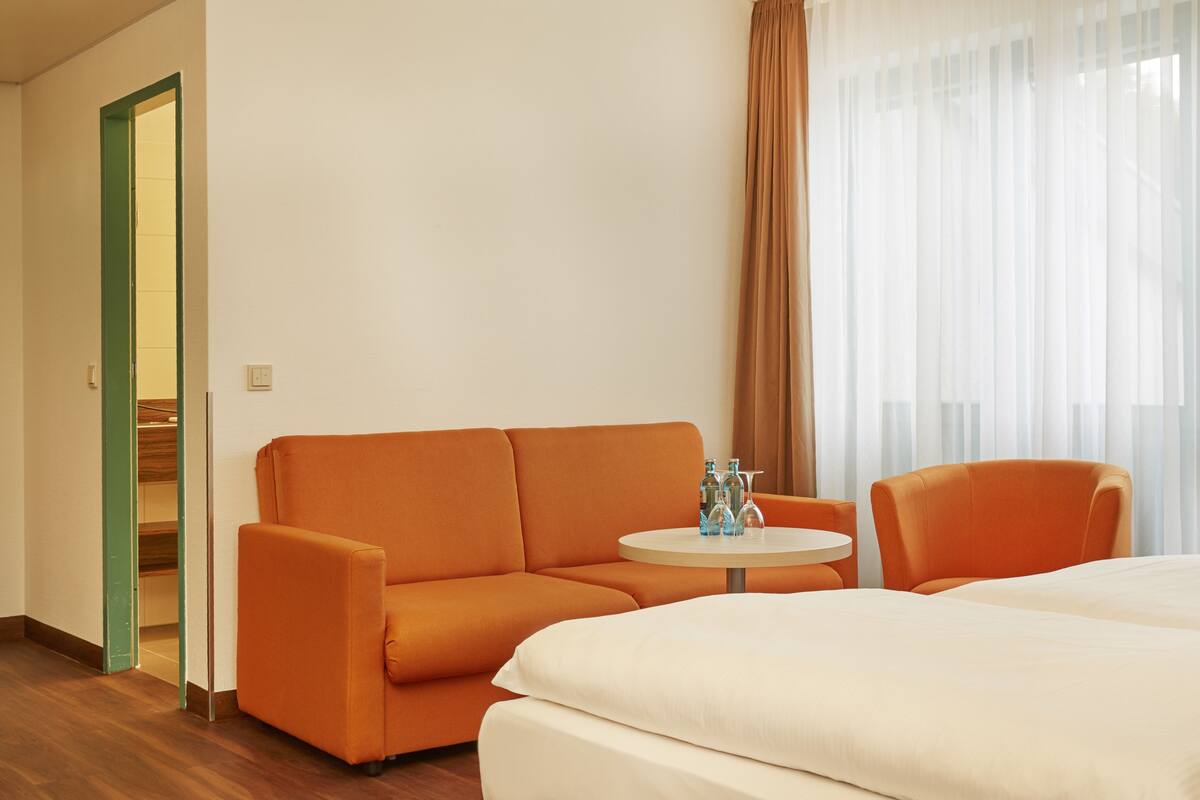 Een hotelkamer met een groot bed, een oranje bank, een klein rond tafeltje met flessen en een raam met dunne gordijnen.