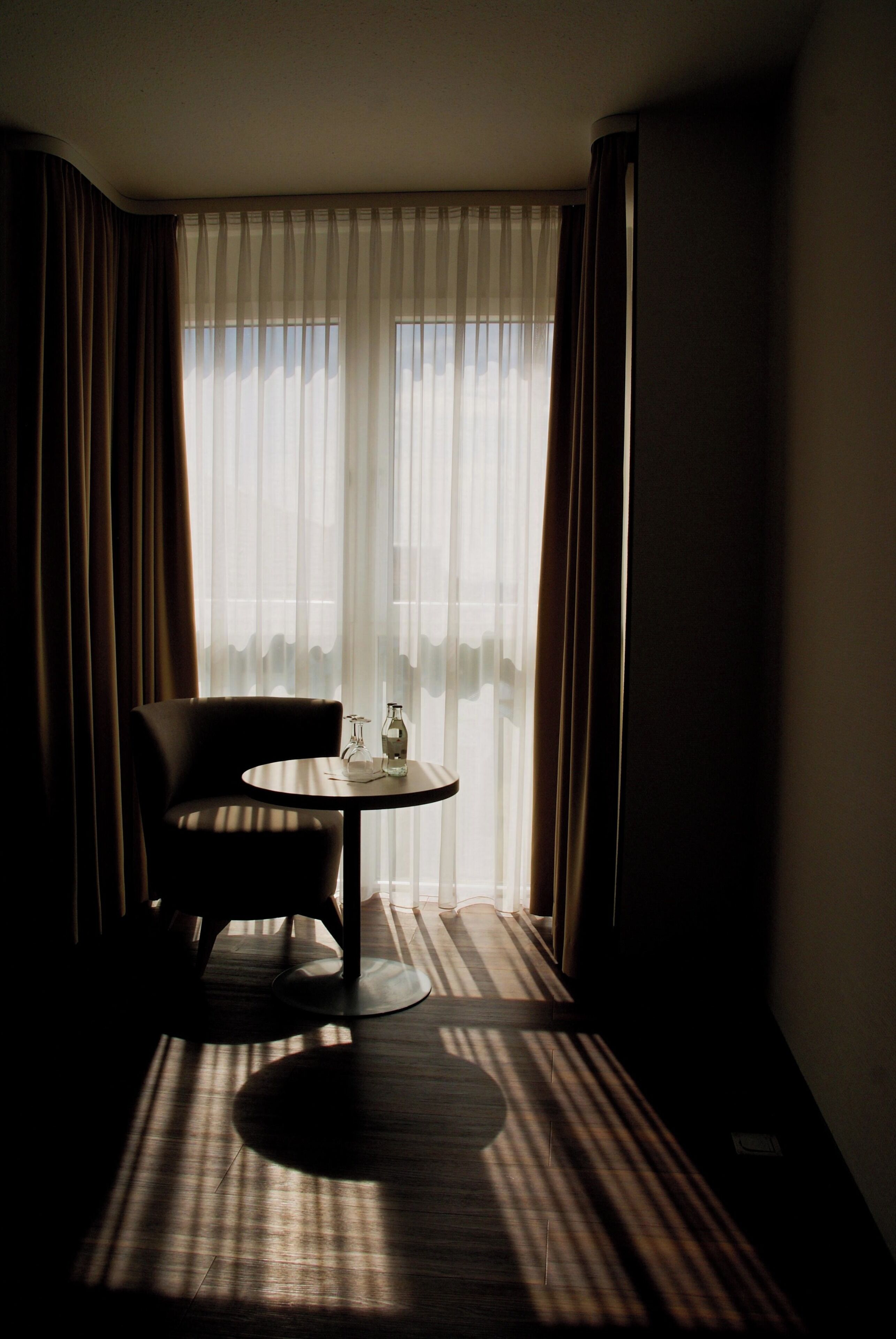 Una habitación de hotel con una silla, una mesita pequeña, una ventana con cortinas ligeras y vista al cielo.