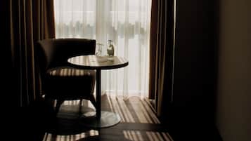 Una habitación de hotel con una silla, una mesita pequeña, una ventana con cortinas ligeras y vista al cielo.