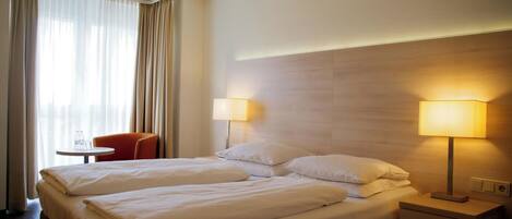 A hotel room with a bed, two bedside lamps, a small table with a chair, and a window with curtains.