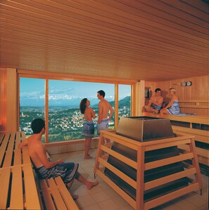 Een sauna met houten banken, een massagetafel en grote ramen met uitzicht op een stad.