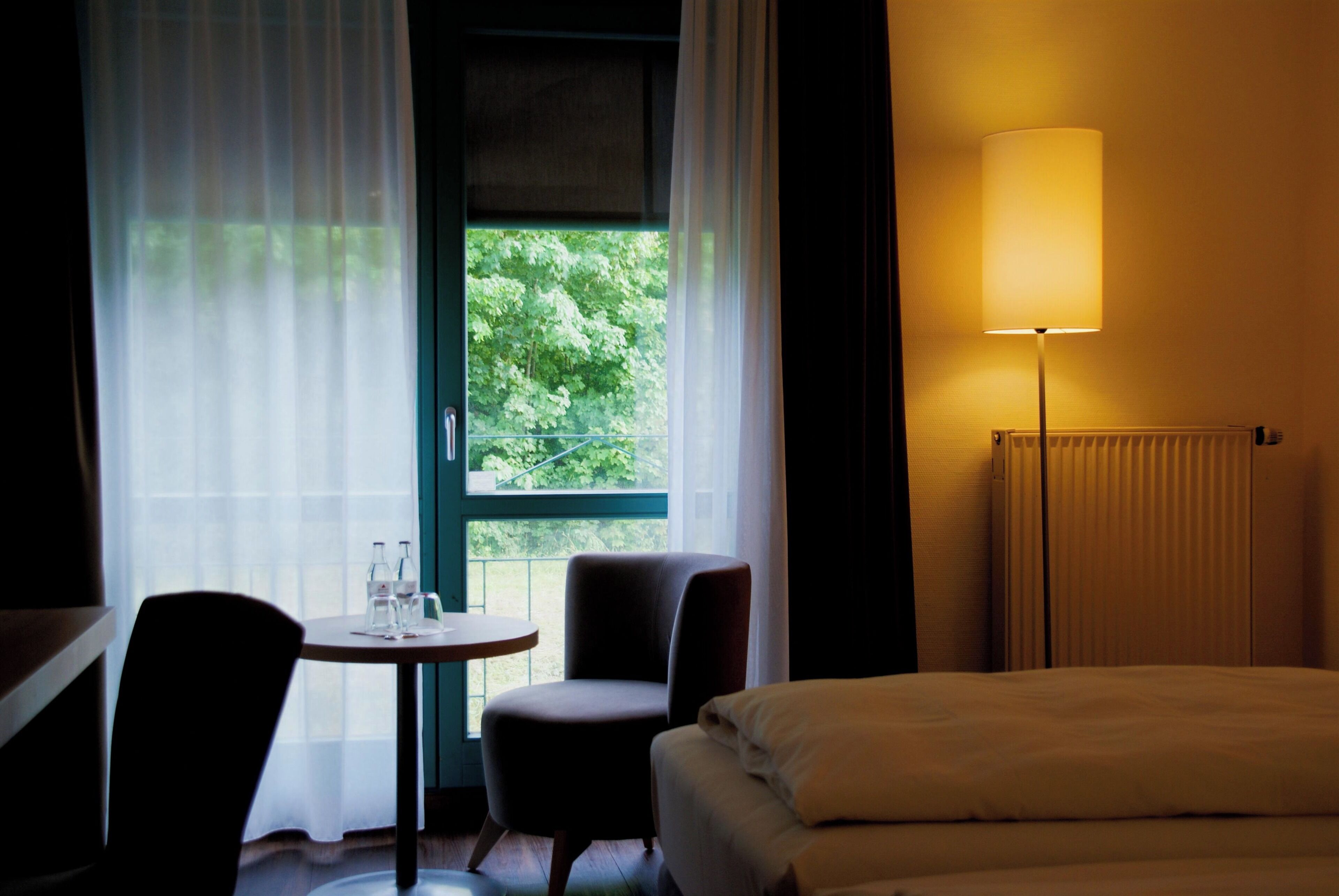 A hotel room with a bed, a table with chairs, a window with curtains, and a standing lamp.