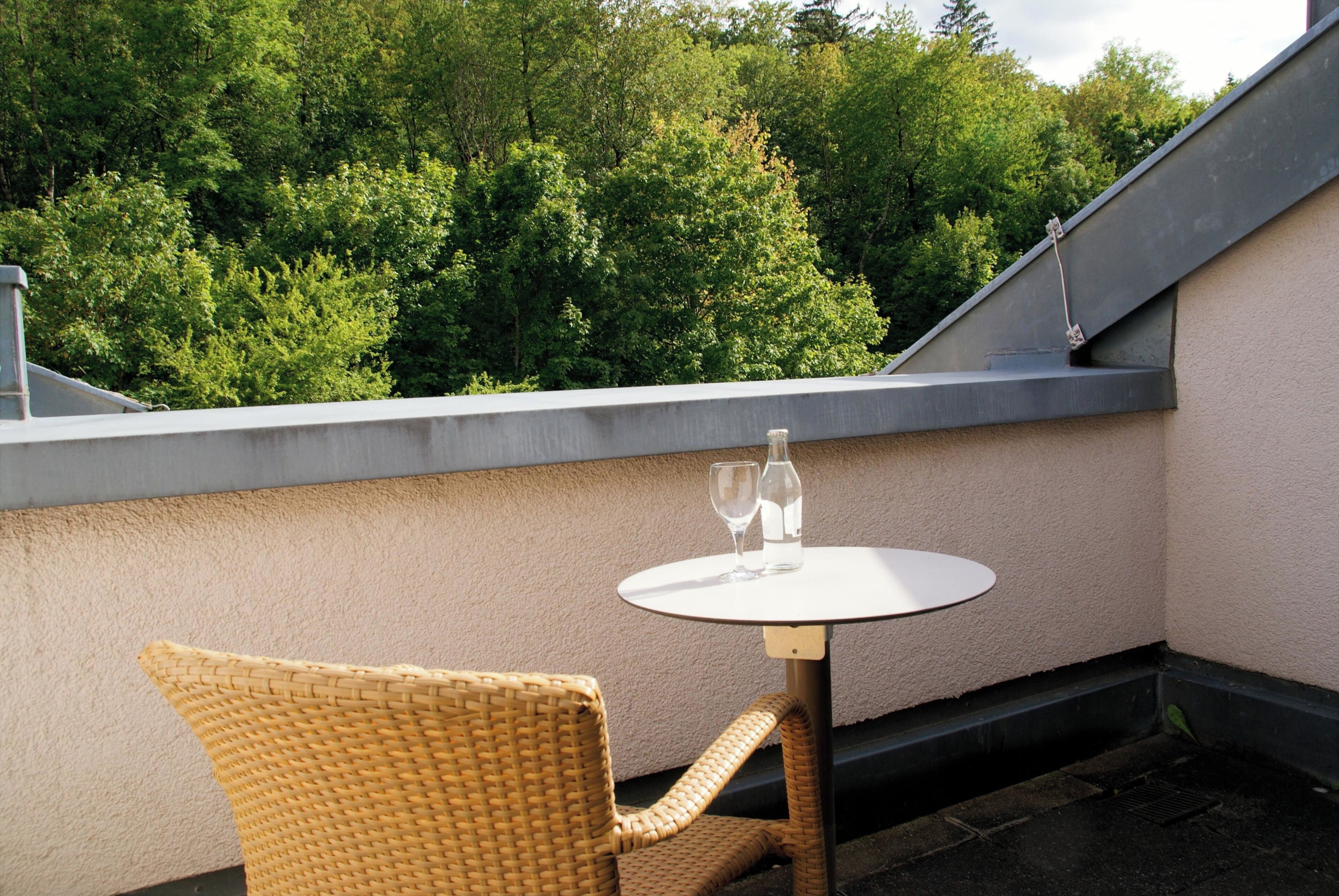 A small outdoor seating area with a round table and wicker chair, overlooking a dense green tree line.