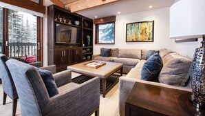 Una sala de estar acogedora con un sofá, sillones, una mesa de centro y un televisor. Se observa una estantería y un cuadro en la pared.