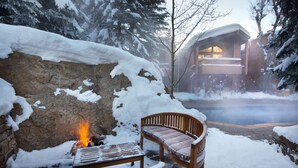 Un banco y una mesa de madera con un pozo de fuego en un espacio exterior nevado, rodeado de árboles cubiertos de nieve y un edificio con balcón.