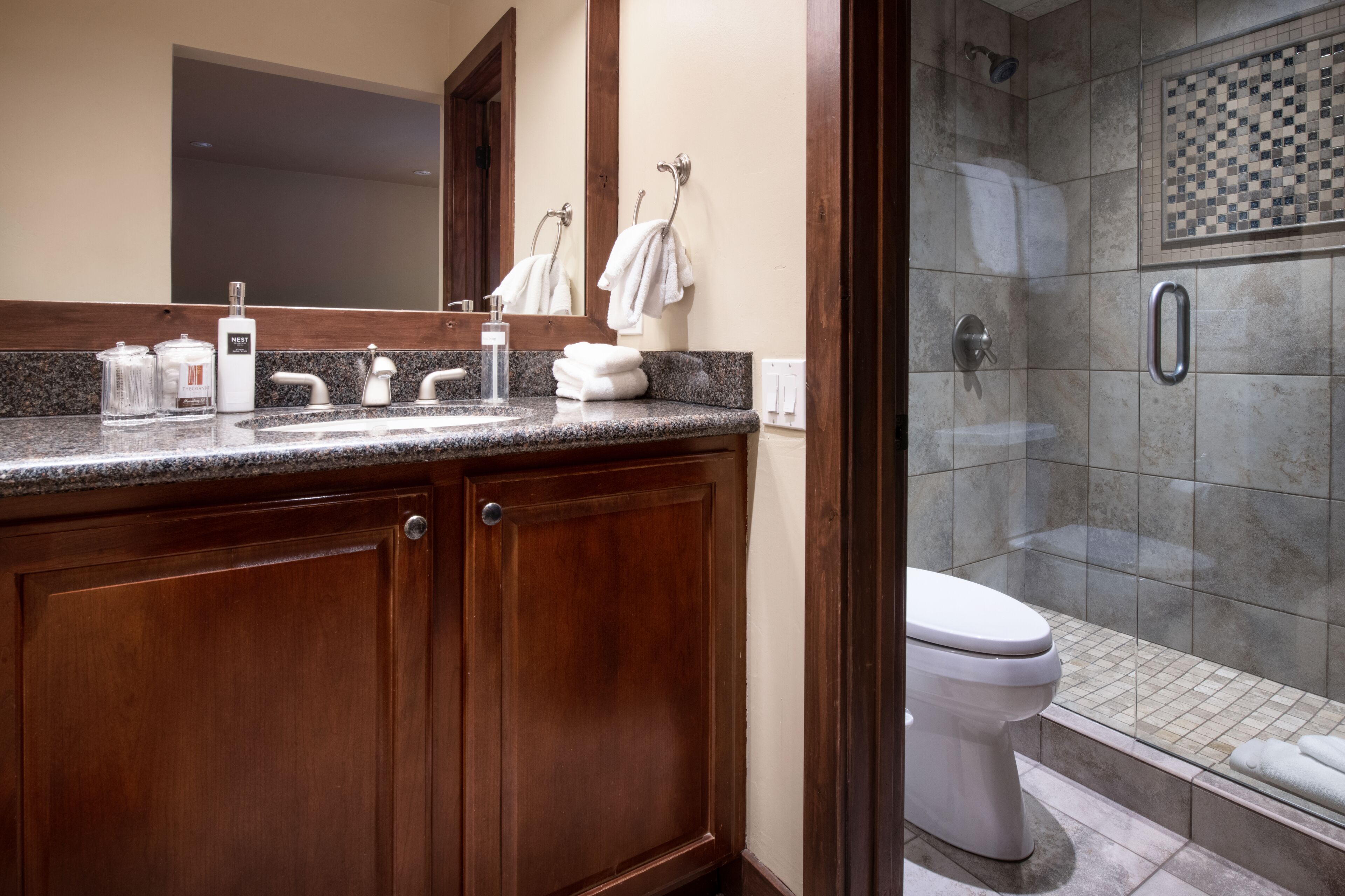 A bathroom with a granite countertop, wooden cabinets, a white toilet, and a shower with a checkered tile wall.