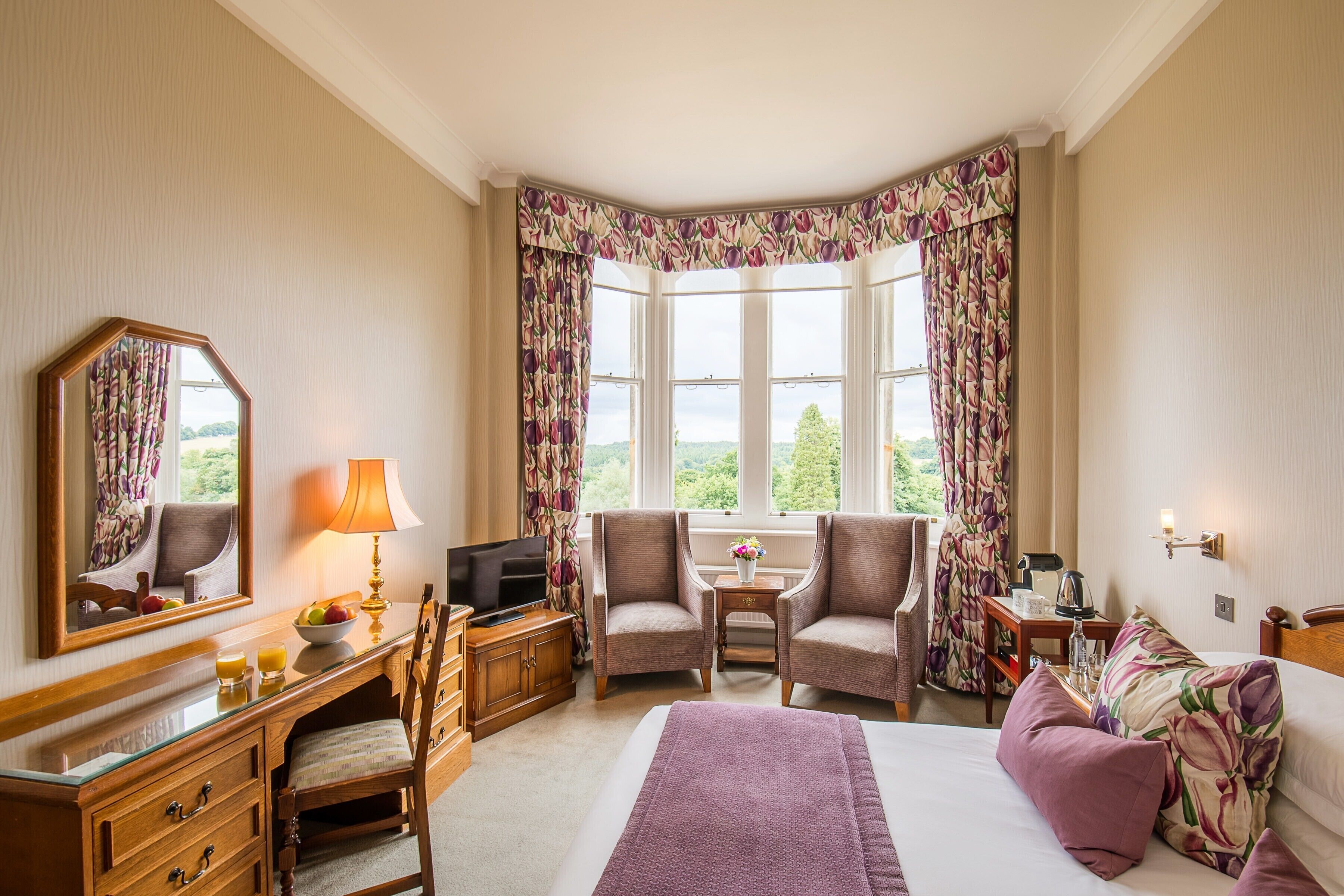 A hotel room with a large bed, two armchairs, a desk with a television, and a window with curtains.
