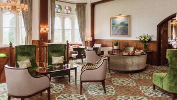 A hotel lobby with green upholstered chairs, a wooden reception desk, and a patterned carpet.
