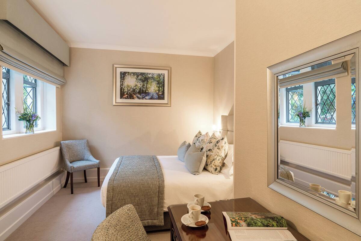 A bedroom with a bed, a chair, a table with cups and a book, a window with a view of greenery, and a framed picture on the wall.