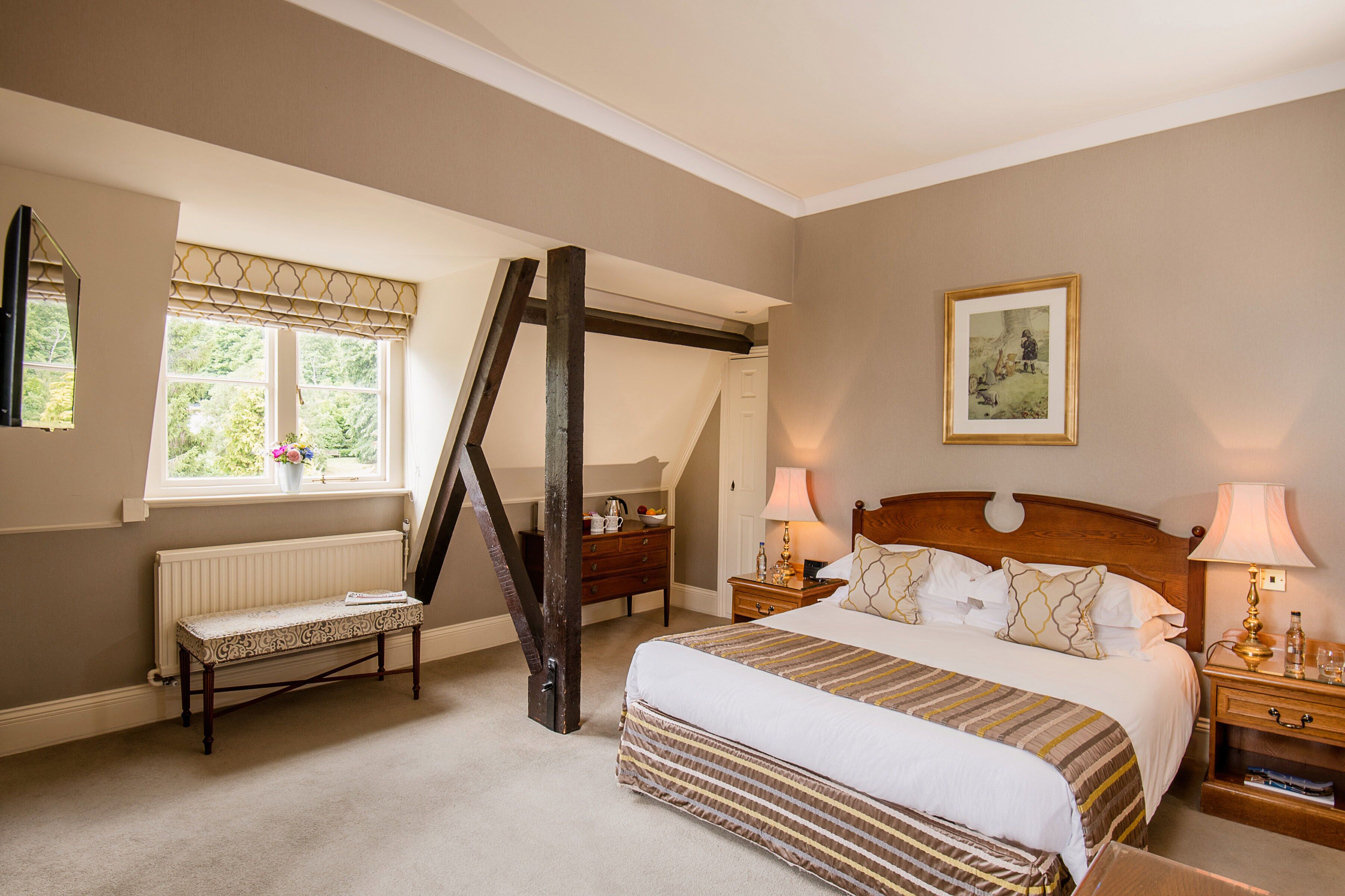A bedroom with a large bed, two bedside tables with lamps, a bench, a radiator, a window with curtains, and a painting on the wall.
