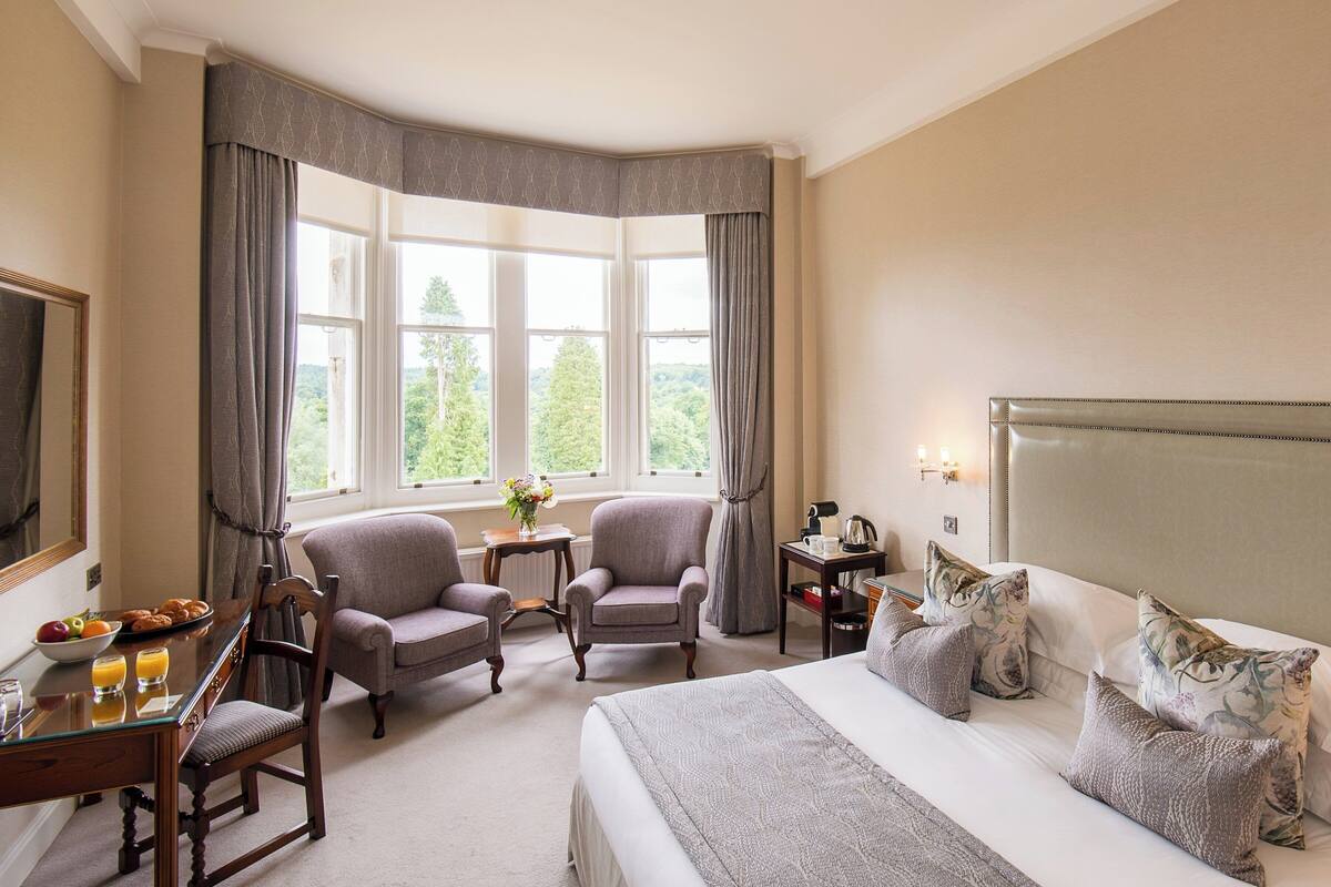 A hotel room with a large bed, two armchairs, a dining table with breakfast, and a view of trees through the windows.