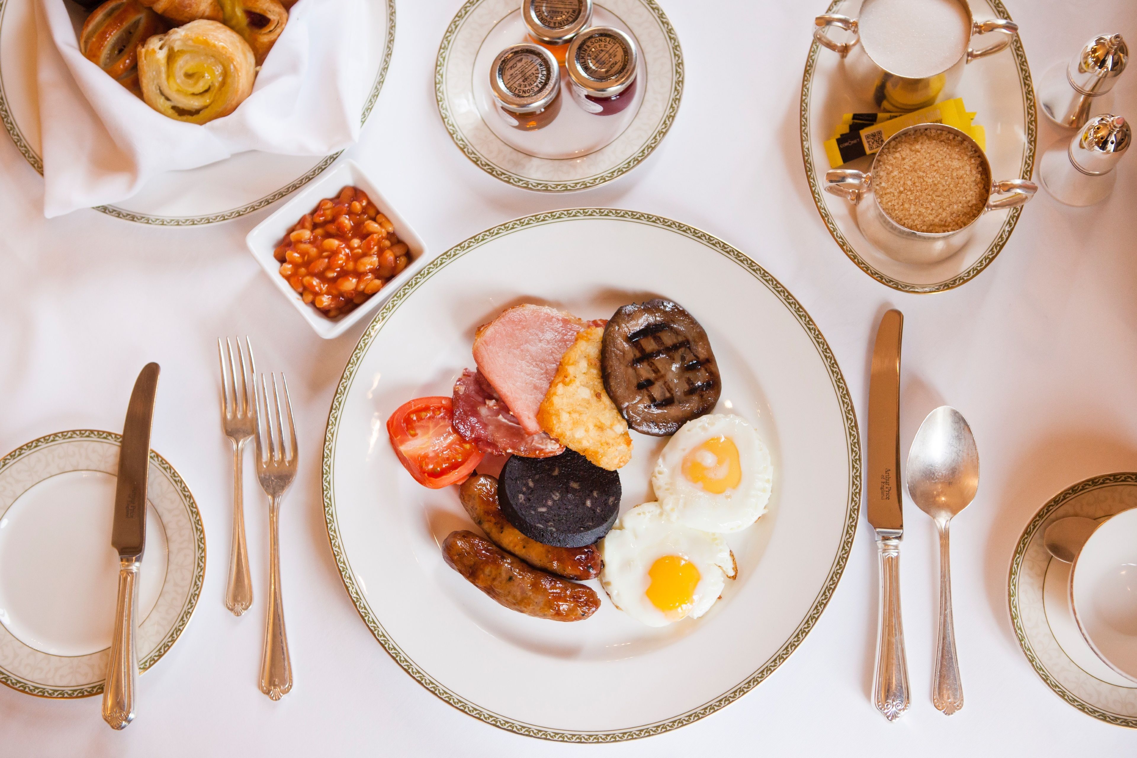 Un desayuno con huevos, salchichas, tomates y morcilla, acompañado de salsas y utensilios.