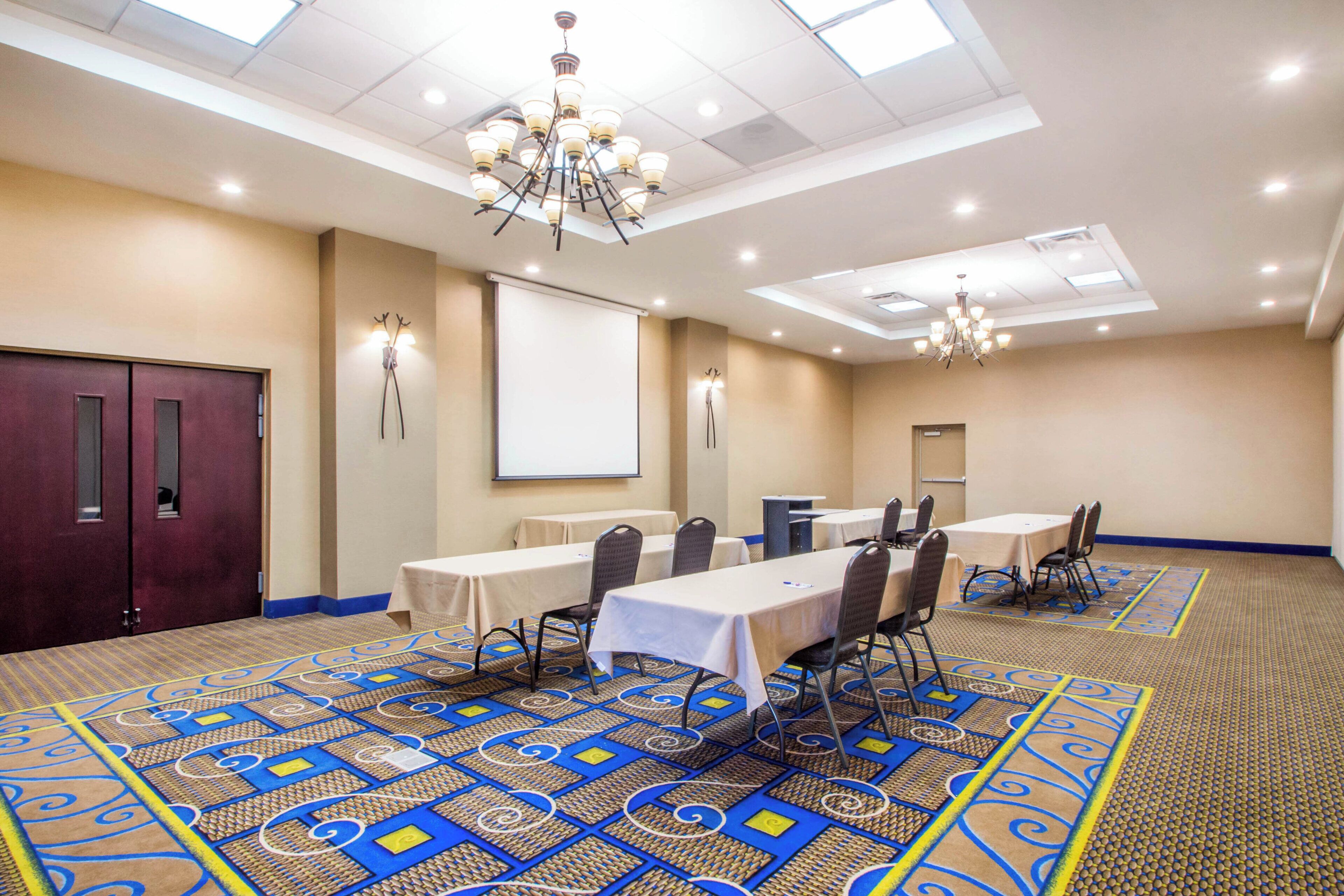 Un salón de conferencias con una pantalla grande, varias mesas, sillas, piso alfombrado con un diseño geométrico y una araña.