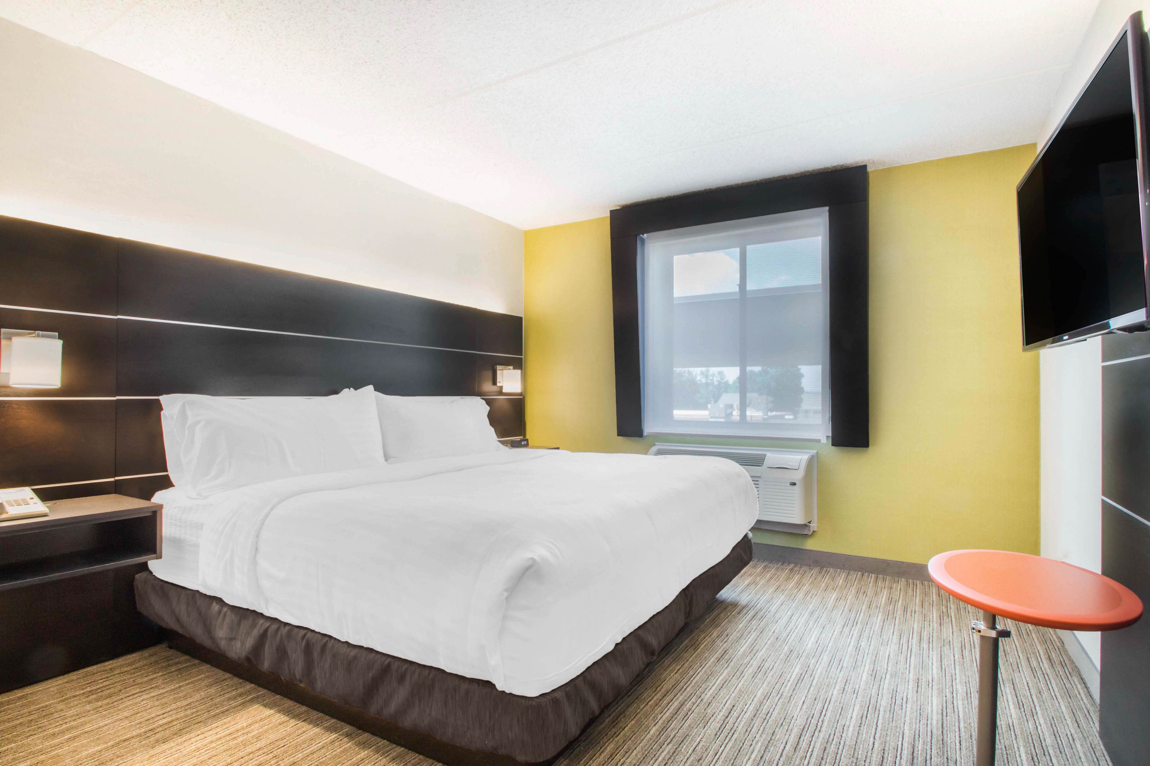 A hotel room with a large bed, a bedside table with a lamp, a flat-screen TV mounted on the wall, and a window with a view of the outside.