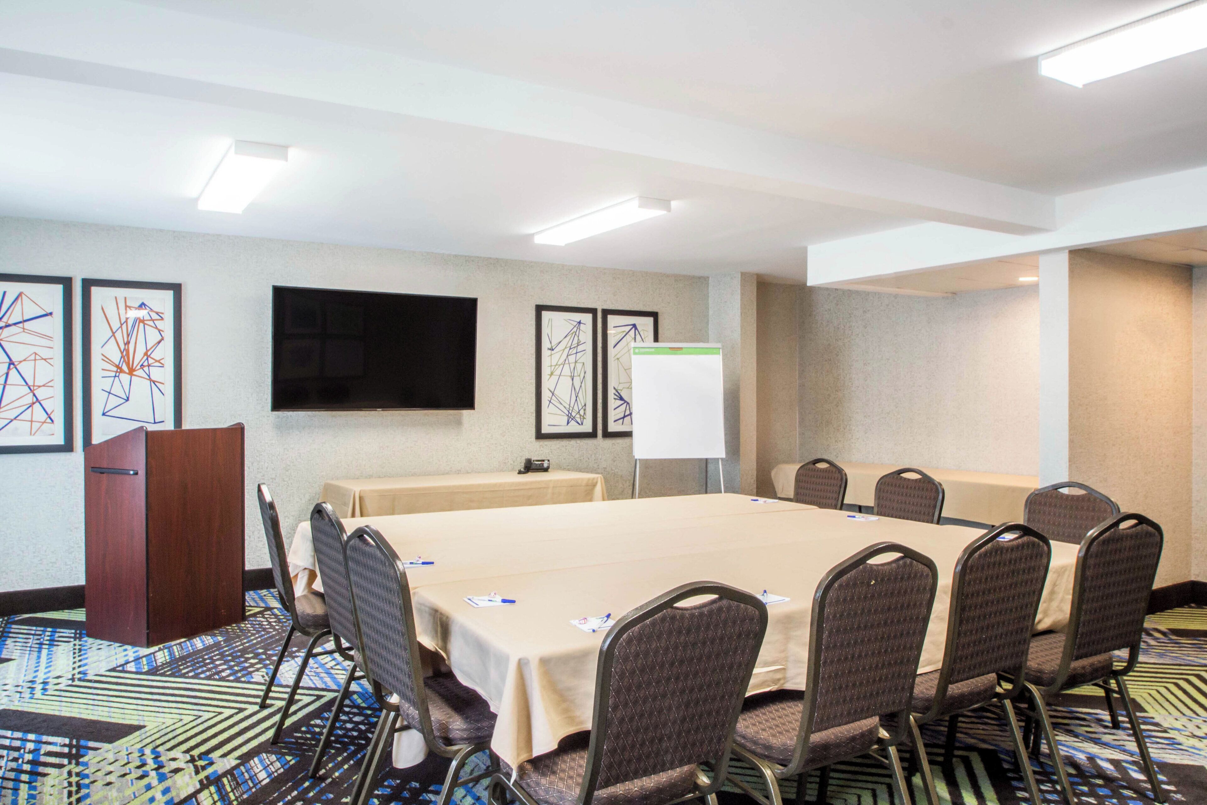 A conference room with a long table, chairs, a podium, a whiteboard, and framed abstract art on the walls.
