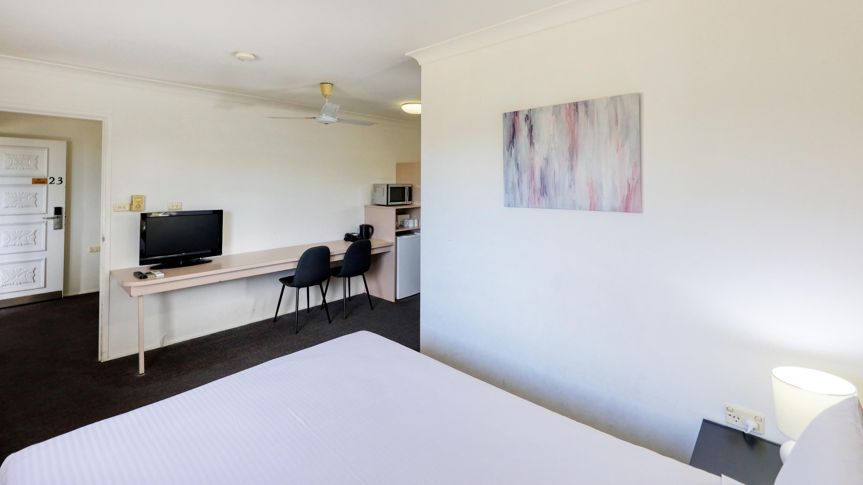 A hotel room with a bed, a desk with a television, two chairs, a microwave, and a painting on the wall.