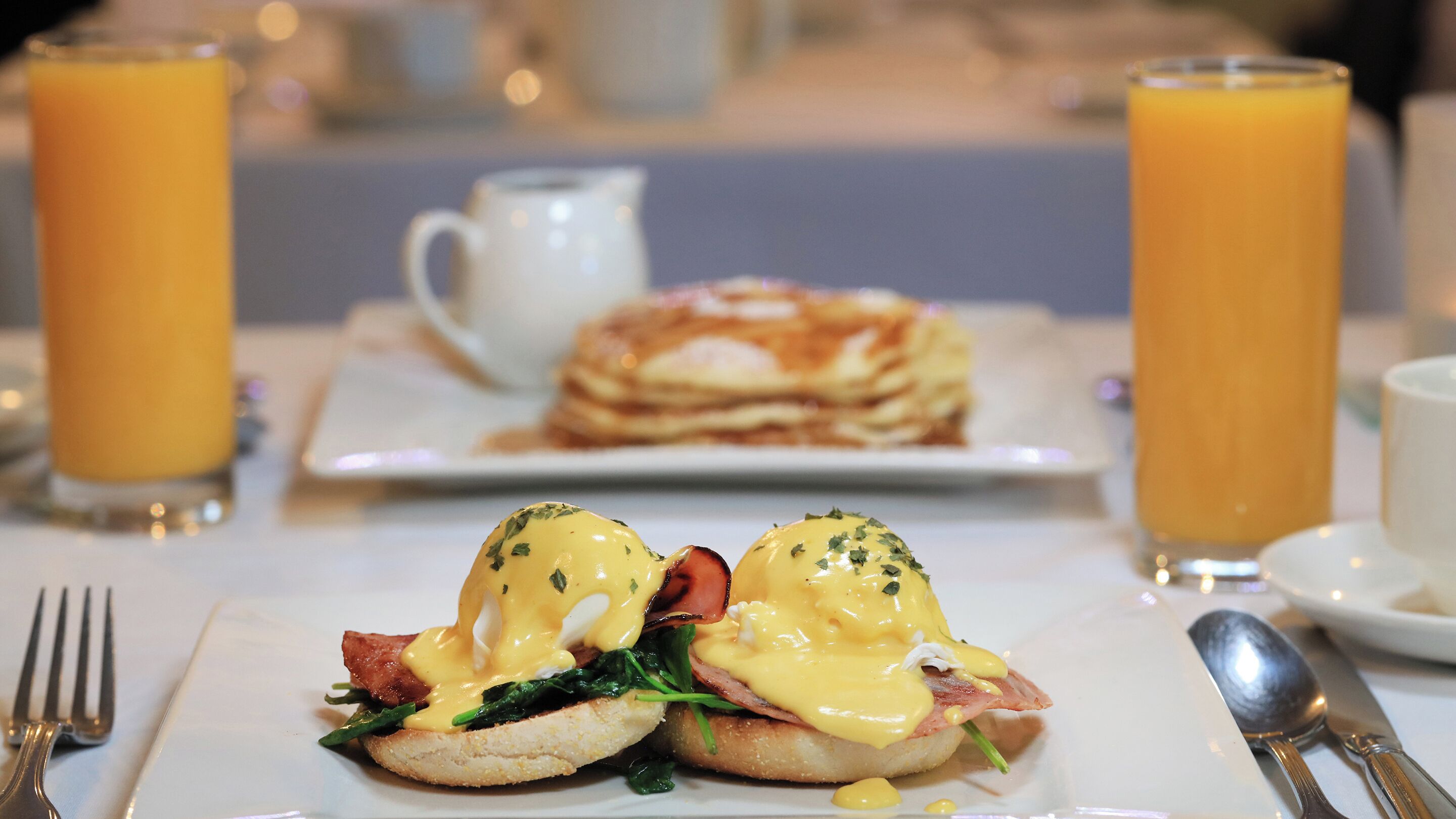 Un desayuno con huevos benedictinos, una taza de café y vasos de jugo de naranja sobre una mesa.