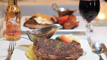 Una mesa de comedor con un filete, puré de papas y vegetales. Hay una botella de vino y una copa de vino tinto sobre la mesa.