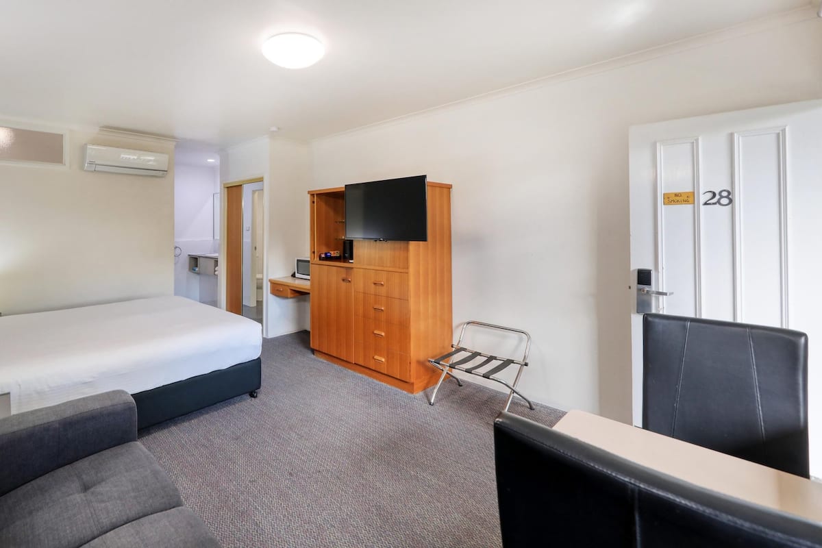 A hotel room with a bed, a desk with a chair, a TV, a wardrobe, and a door with a number.