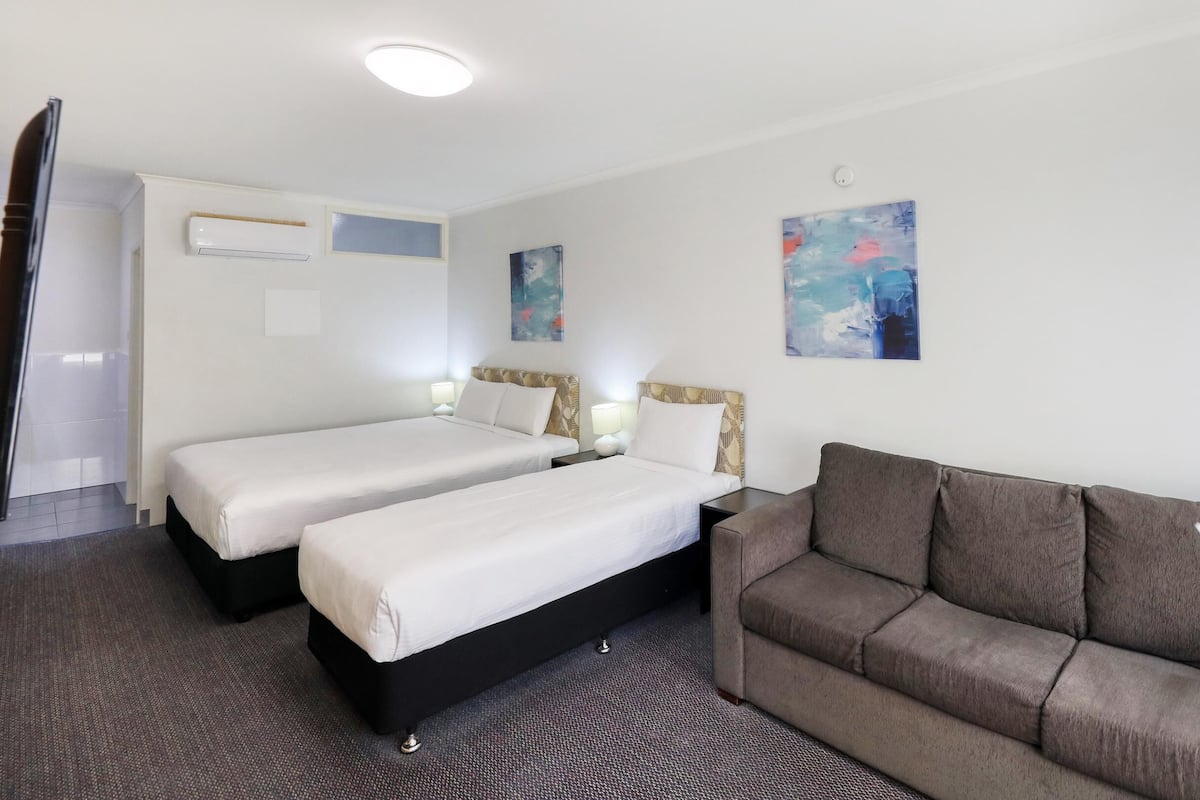A hotel room with two single beds, a sofa, a flat-screen TV, an air conditioning unit, and two paintings on the walls.