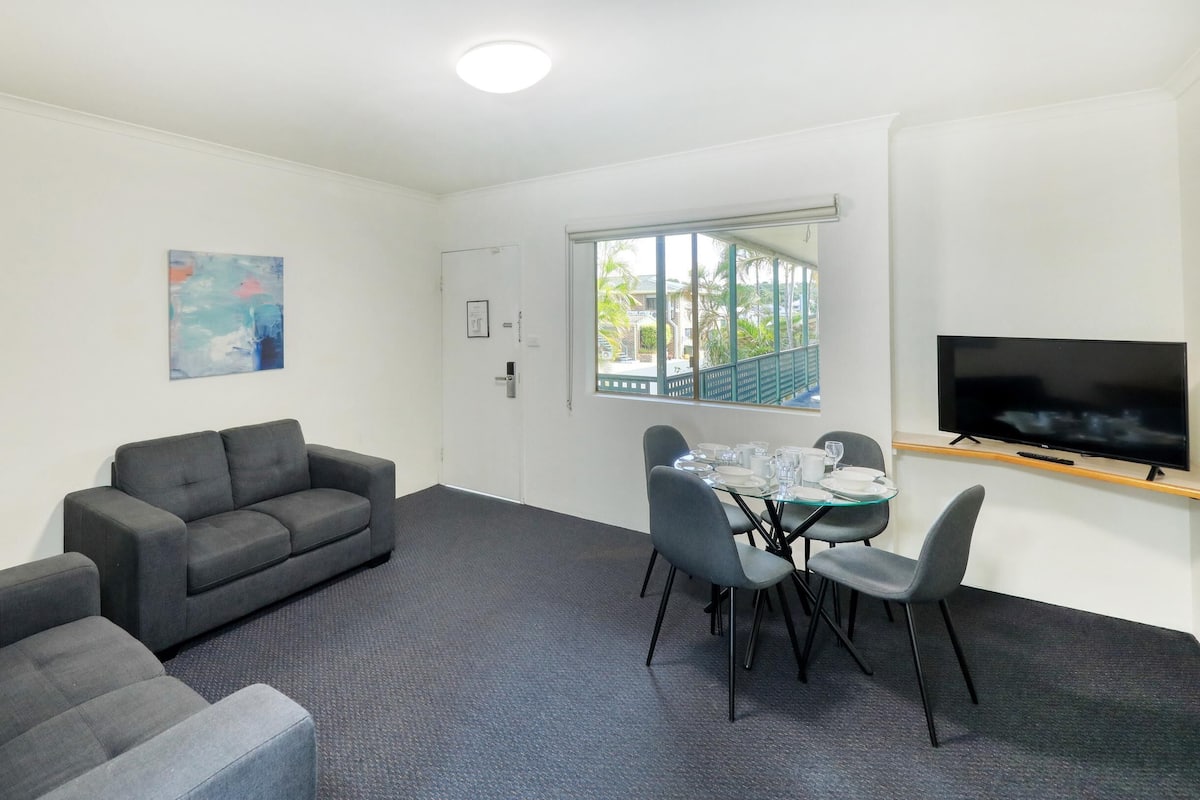 A modern, well-lit room with a grey sofa, a dining table set for four, a flat-screen TV mounted on the wall, and a window offering a view of greenery outside.