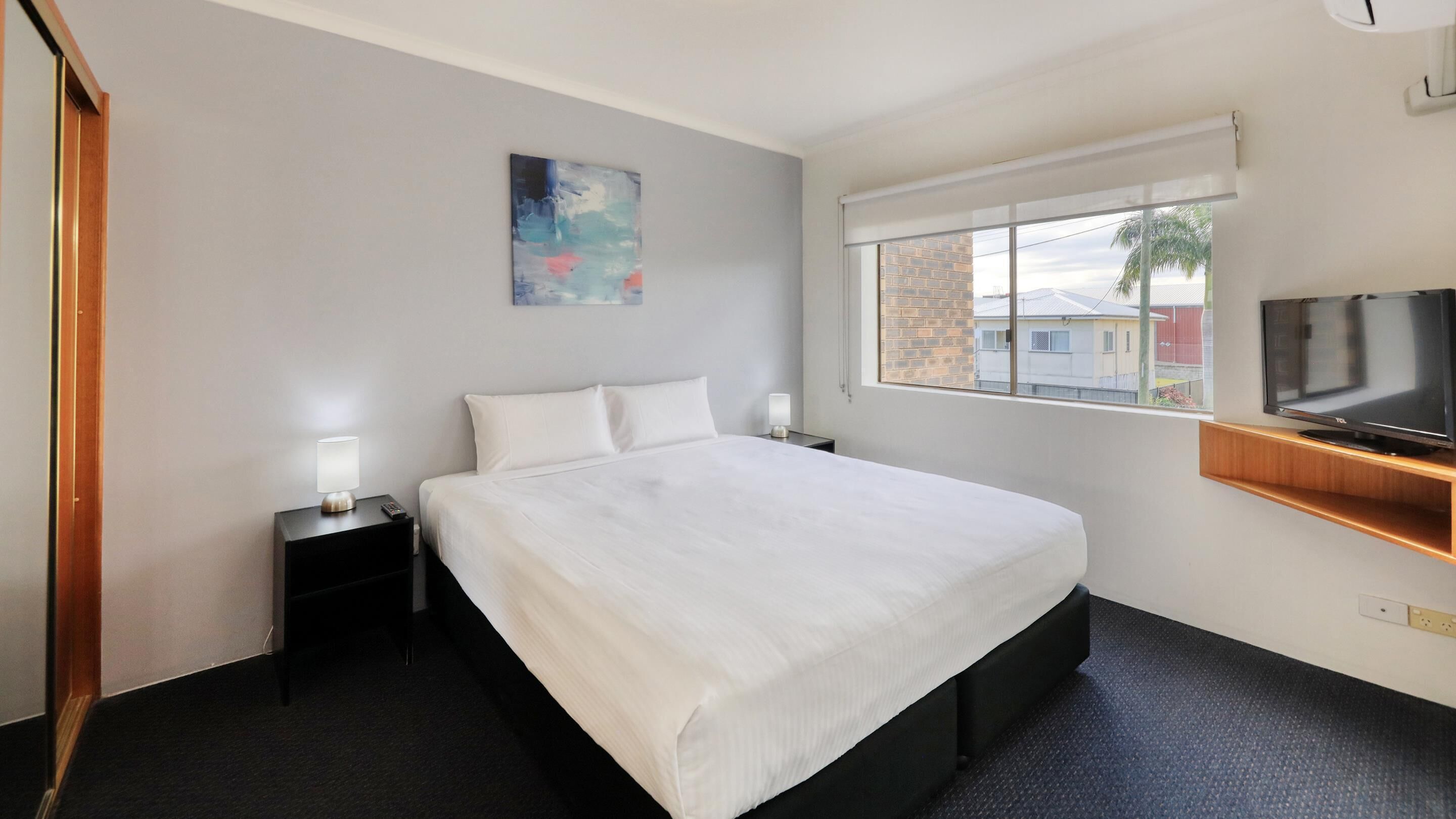 A hotel room with a large bed, a TV on a wooden shelf, a painting on the wall, and a window with a view of buildings.