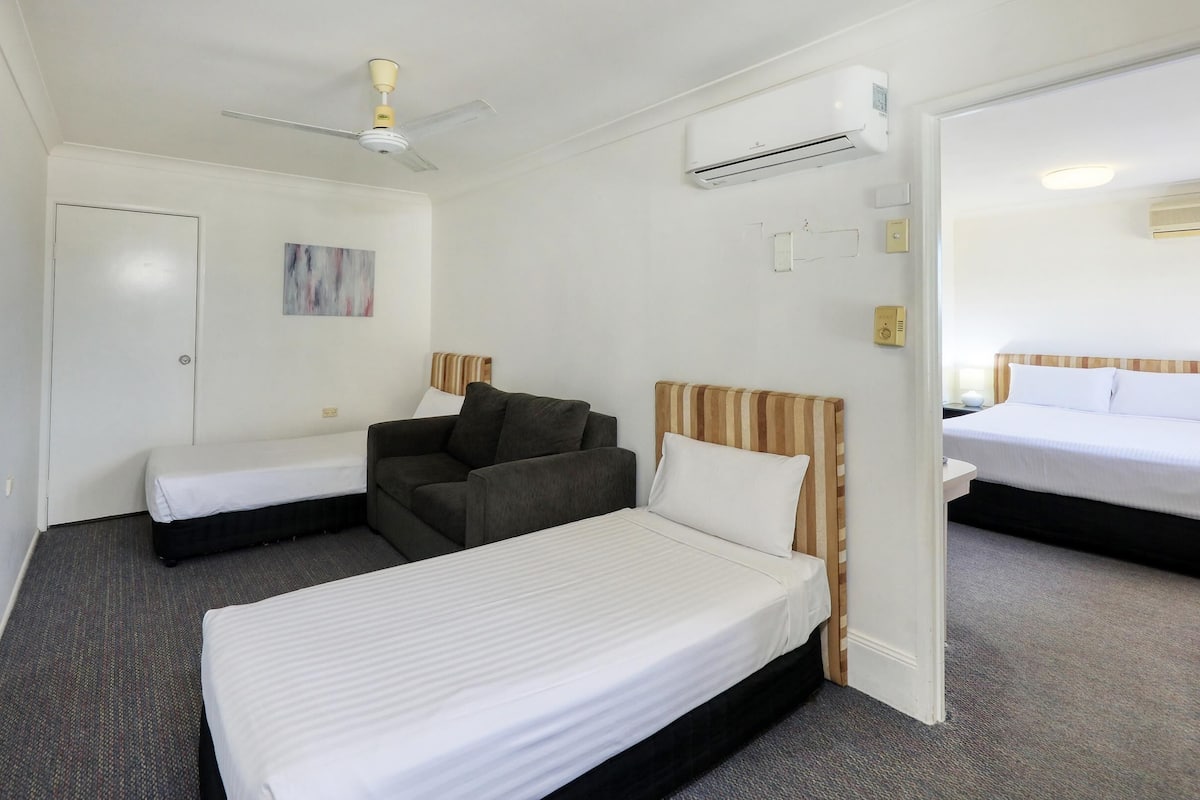 A hotel room with two single beds, a sofa, a ceiling fan, and an air conditioning unit.