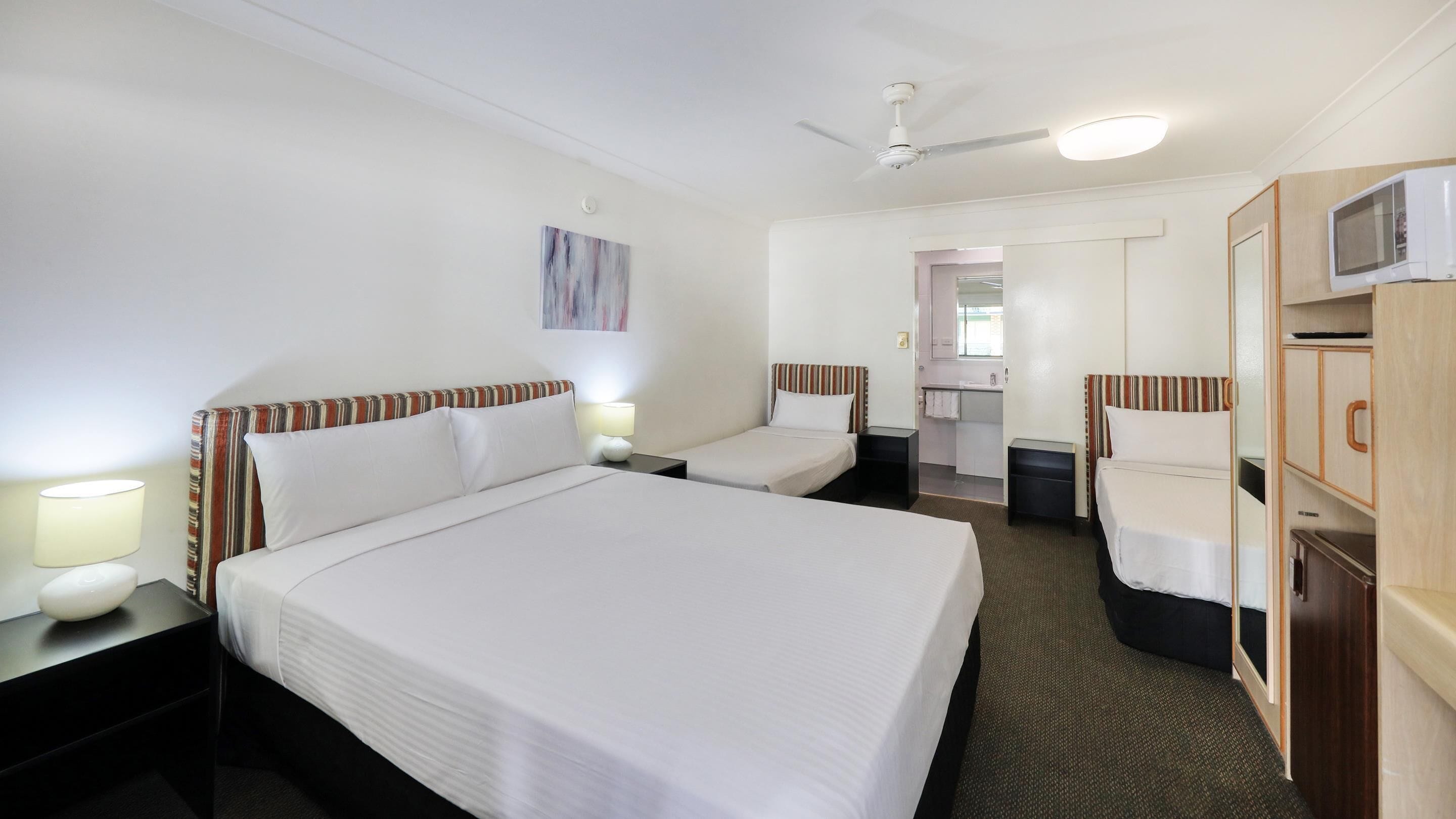 A hotel room with a large bed, two smaller beds, a desk, a wardrobe, a ceiling fan, and a bathroom visible through an open door.