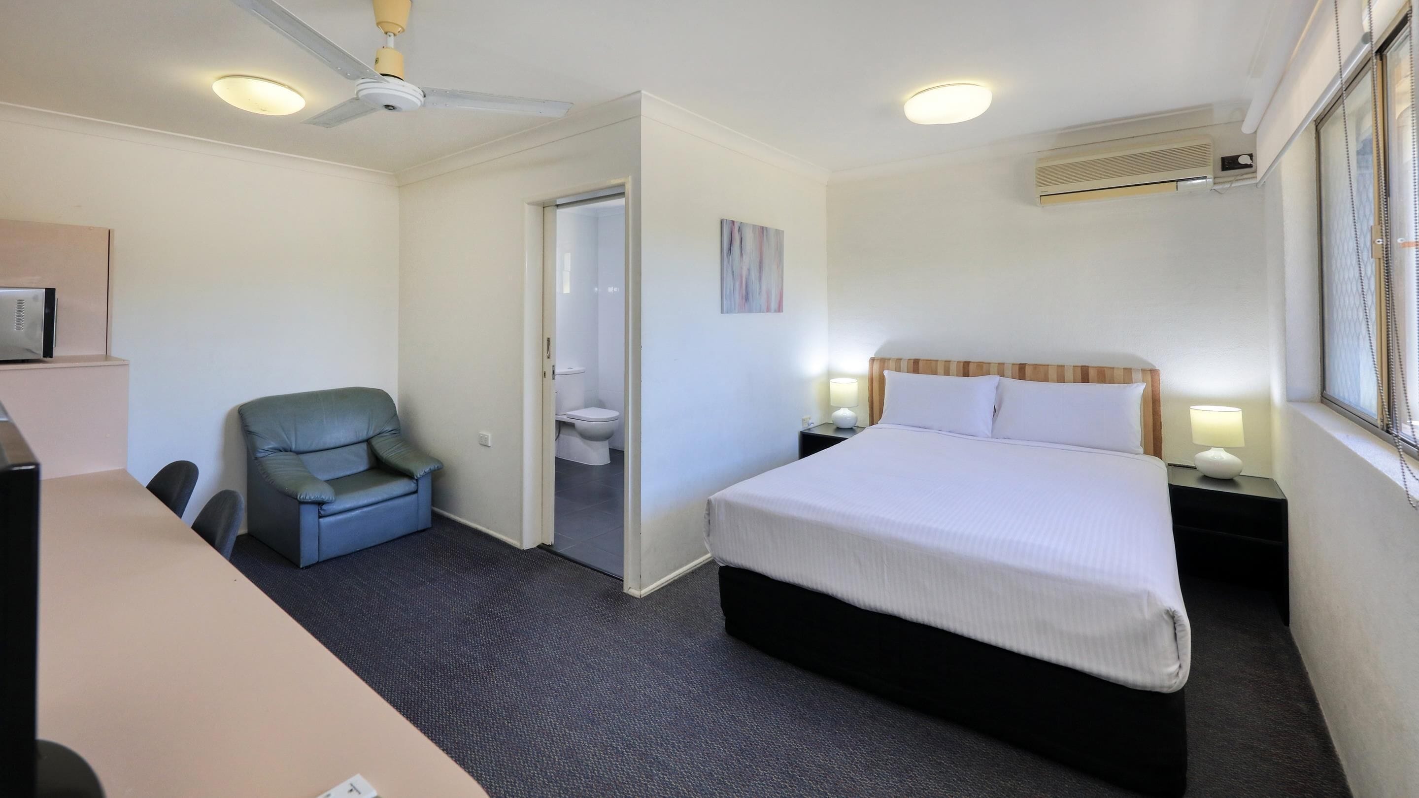 A hotel room with a bed, a desk, a chair, a toilet, and a ceiling fan.