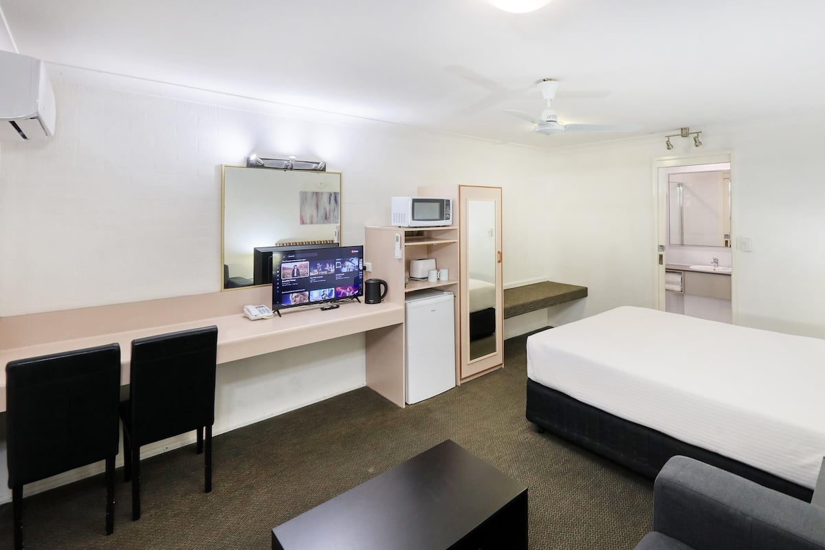 A hotel room with a bed, desk, chairs, a microwave, a coffee maker, a wardrobe, and a ceiling fan.
