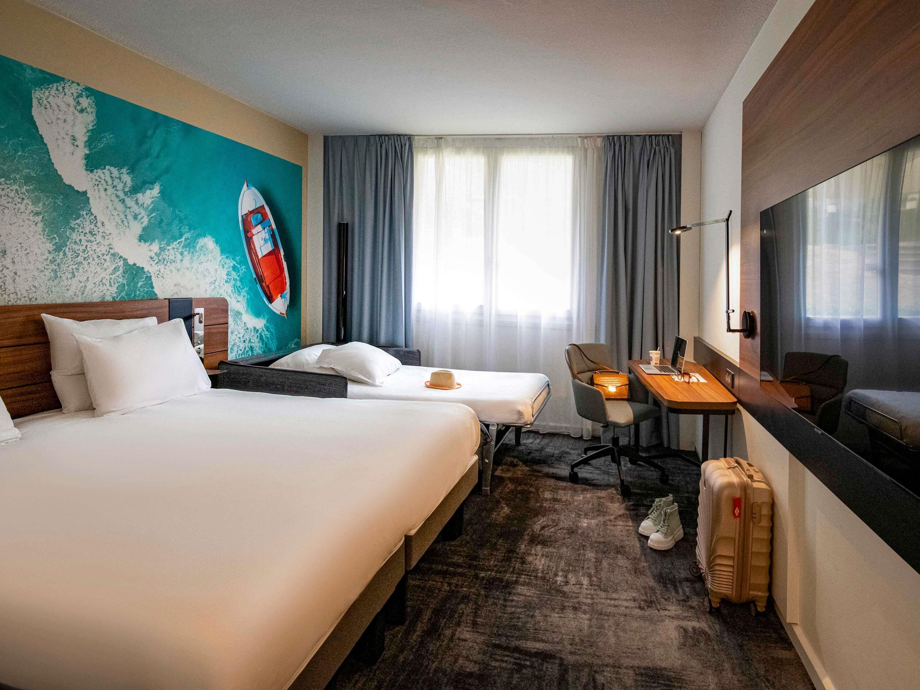 Une chambre d’hôtel avec un grand lit, un bureau, une chaise, une valise et un mur décoré d’une fresque représentant une planche de surf.