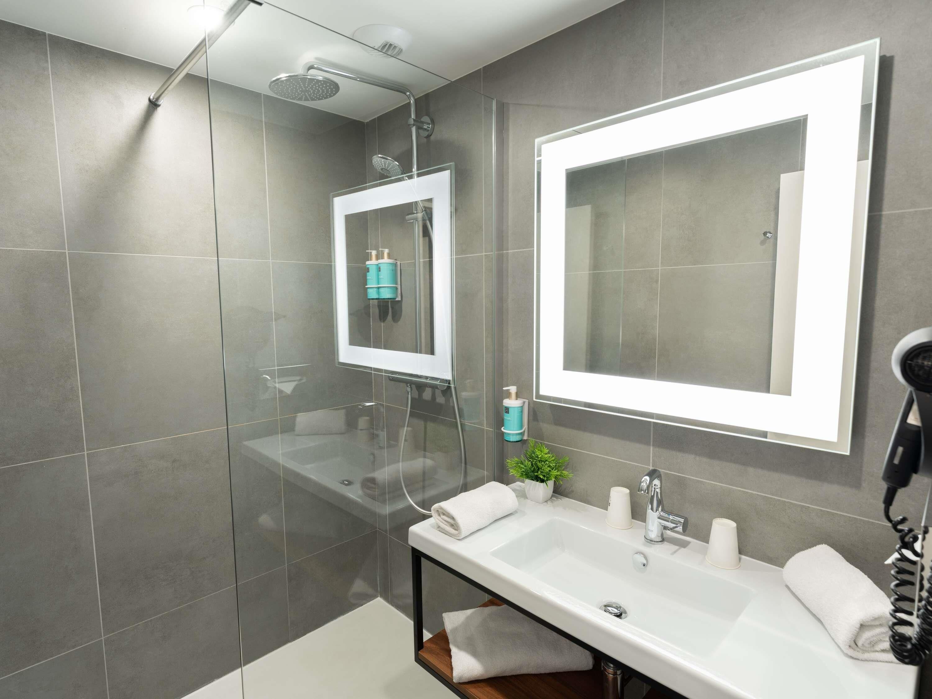 Un baño moderno con ducha de vidrio, un espejo grande, un lavabo blanco y un secador de pelo.