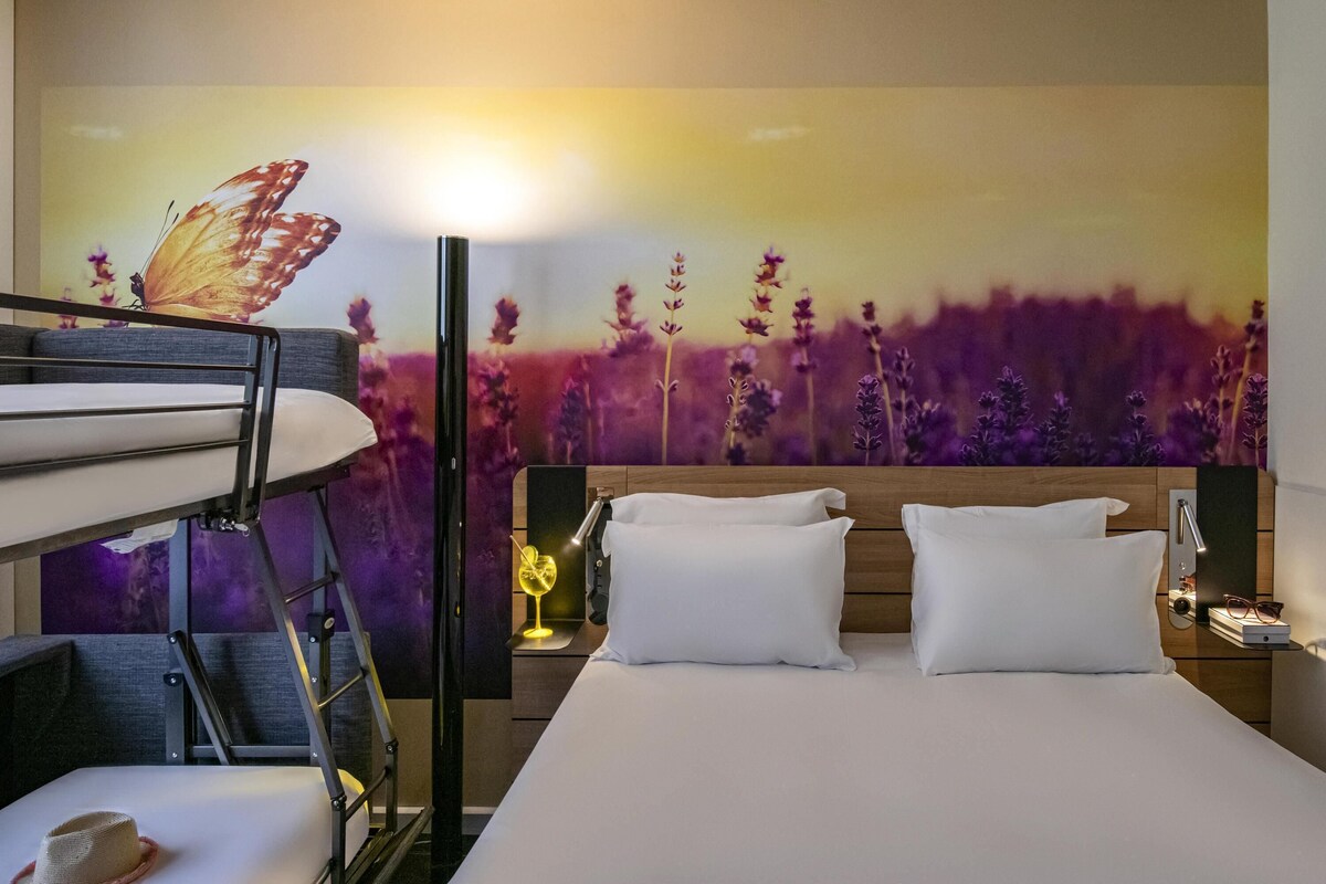 Una camera d'albergo con letti a castello, un letto con lenzuola bianche, un comodino con una lampada e un affresco floreale con una farfalla.
