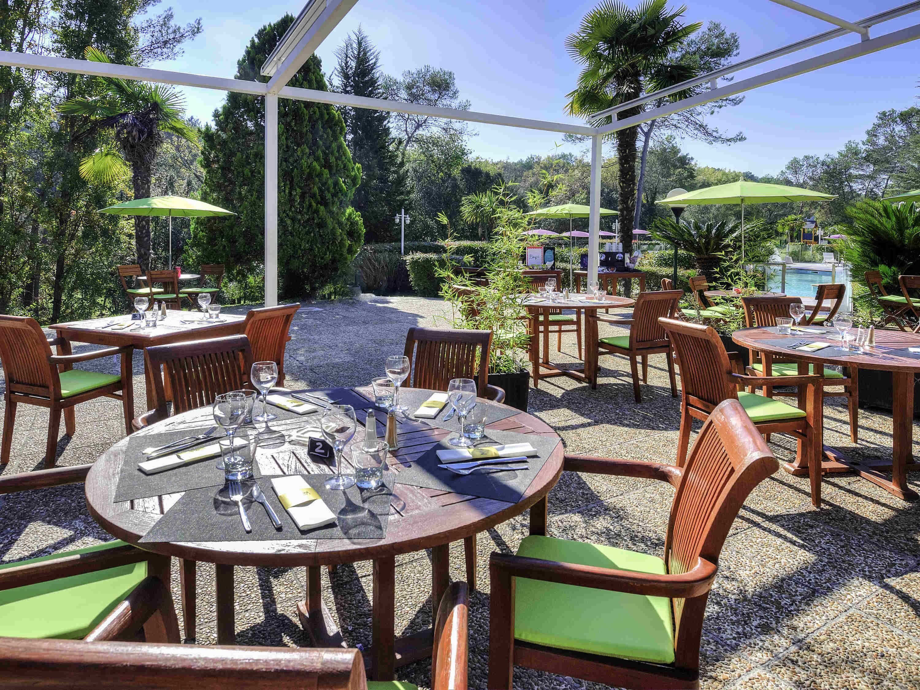 Een terras buiten met ronde houten tafels en stoelen, waarvan sommige voorzien zijn van groene kussens.