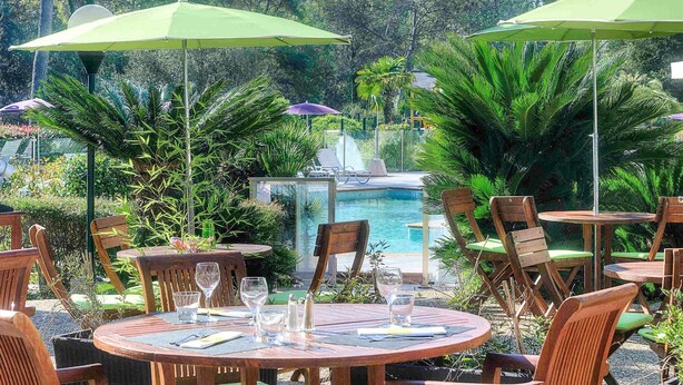 Un espace aménagé au bord de la piscine, avec des tables et des chaises rondes en bois, des parasols verts et une végétation luxuriante.
