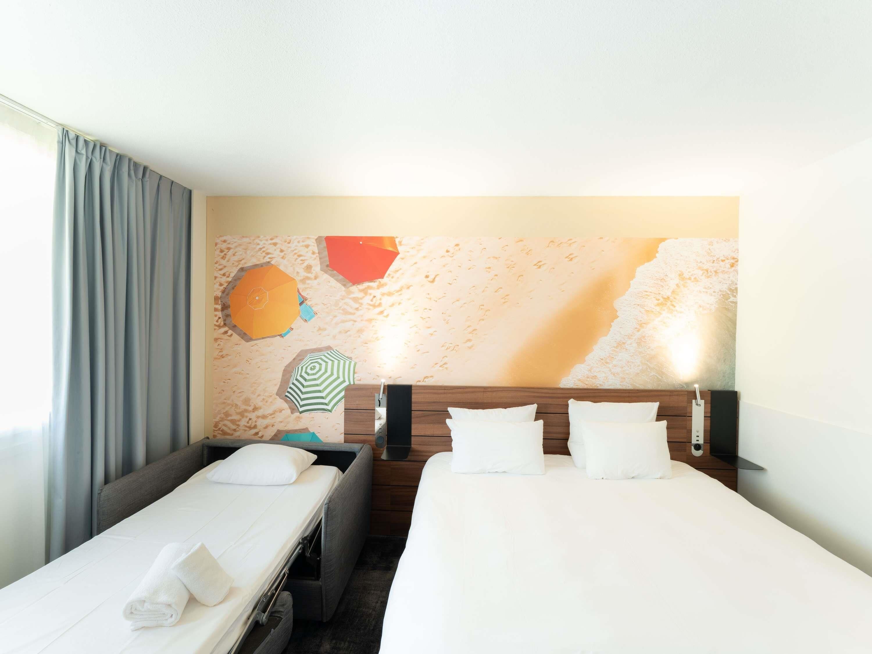 Une chambre d’hôtel avec deux lits, une grande fresque murale sur le thème de la plage et une fenêtre avec des rideaux.