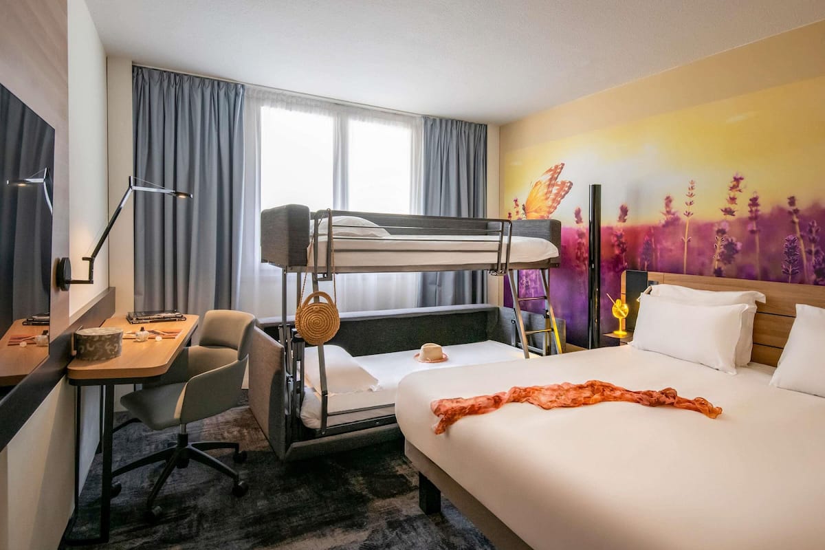 Een hotelkamer met een stapelbed, een bureau met stoel, een nachtkastje en een groot raam.