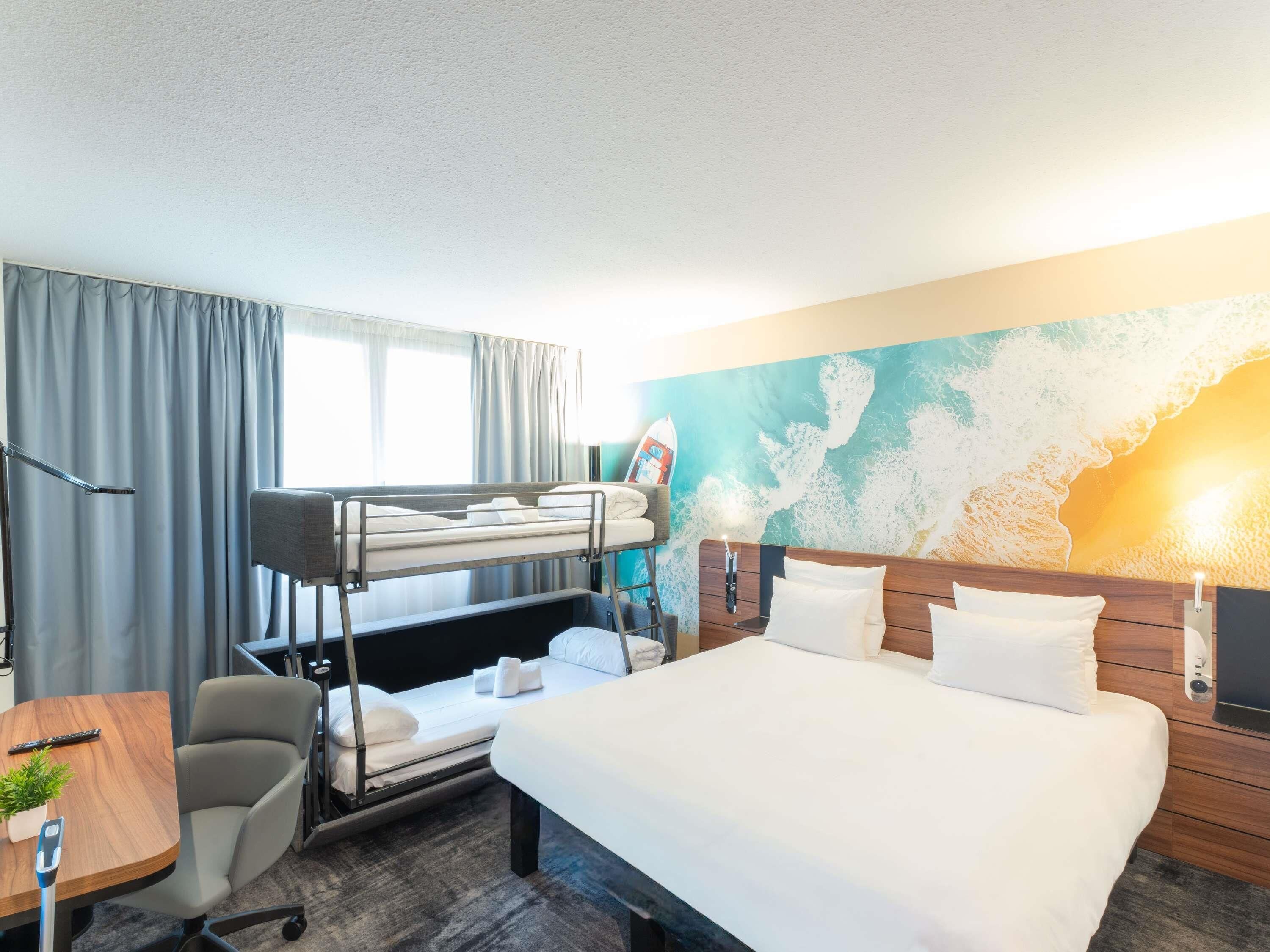 A hotel room with a bunk bed, a desk with a chair, a bed with a wooden headboard, and a large wall mural.