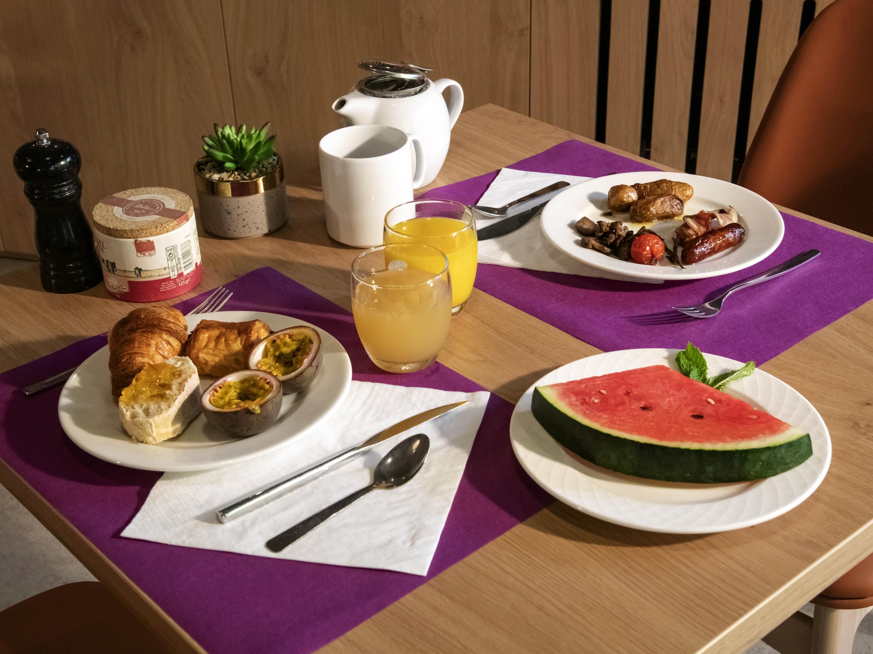 Una mesa de desayuno con platos de comida, una jarra de jugo, una tetera y una pequeña planta.